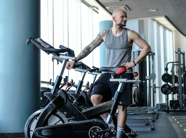 Athletischer Mann mit Tätowierung auf der Hand, der neben einem Heimtrainer im modernen Fitnessclub steht