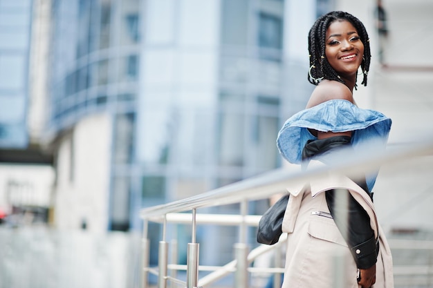 Attraktive afrikanisch-amerikanische Frau mit Dreads in Jacke posierte in der Nähe von Geländern vor einem modernen mehrstöckigen Gebäude