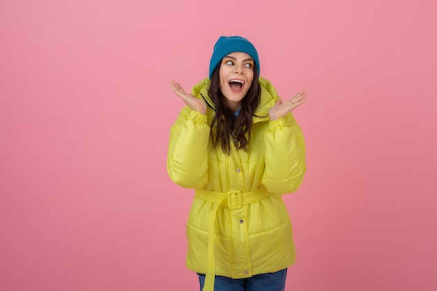 Attraktive aktive Frau, die auf rosa Wand in der bunten Winter-Daunenjacke der leuchtend gelben Farbe aufwirft, lächelnder Spaß, Modetrend des warmen Mantels, verrückter schockierter überraschter Gesichtsausdruck