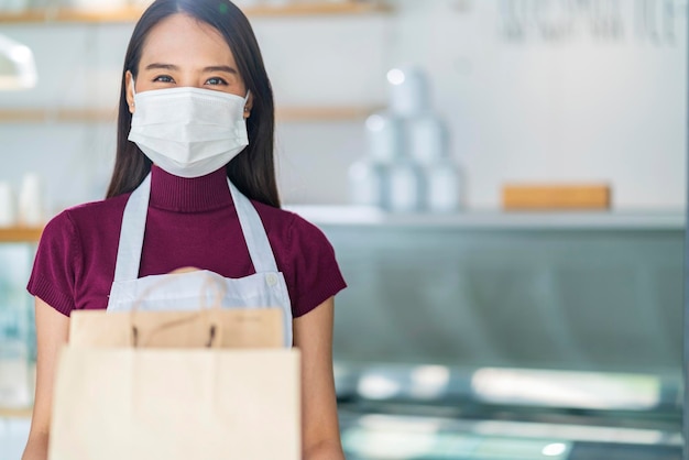 Attraktive asiatische Café-Arbeiterin trägt Gesichtsmaske und Handschuhe und gibt der Kundenkellnerin, die die Bestellung zum Mitnehmen hält, im Café-Hauseingang einen neuen normalen Lebensstil