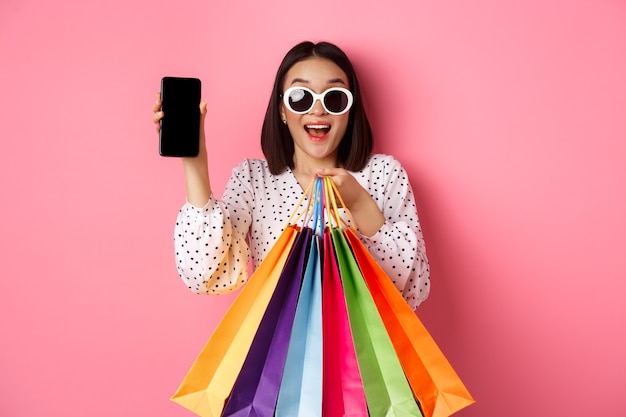 Attraktive asiatische Frau, die Smartphone-App und Einkaufstaschen zeigt, die online über den Anwendungsstand...