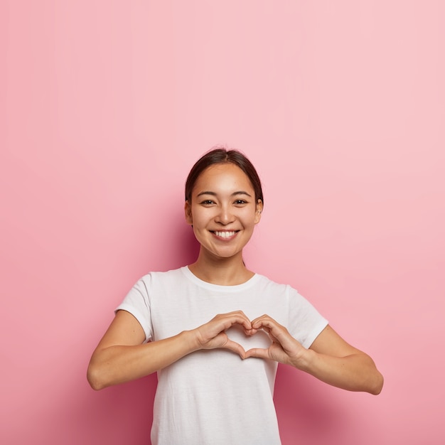 Attraktive asiatische Frau macht Herzform Geste, drückt Liebe aus, sagt sei mein Valentinstag, lächelt positiv, trägt weißes Outfit, posiert gegen rosa Wand mit leerem Raum. Körpersprachenkonzept