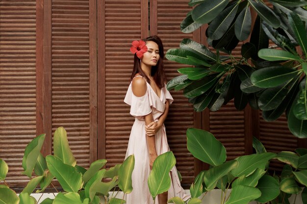 Attraktive asiatische Frau mit Hibiskusblume in den Haaren, die im tropischen Garten aufwerfen.