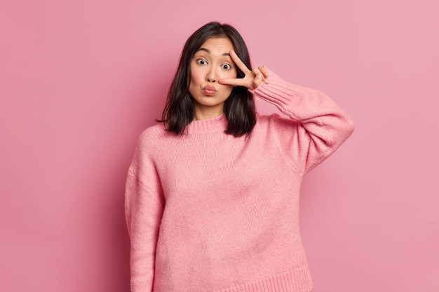 Attraktive asiatische Frau zeigt Siegeszeichen über Auge hält Lippen abgerundet macht Disco-Geste hat Spaß drinnen trägt lässigen Pullover