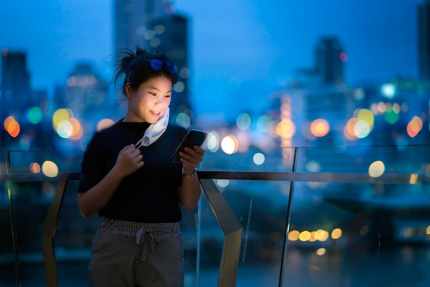 Attraktive asiatische Frauen tragen Schutzmaske Hand verwenden Smartphone-Technologie Kommunikation mit Hintergrund von Bokeh Light Urban Building Hintergrund Geschäftsideen Konzept