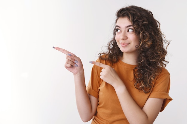 Attraktive begeisterte armenische junge Frau lockiges Haar, die neugierig auf die linke Zeigefinger zeigt, fantastische Leerraumwerbung, die freudig grinst, Produkt einführt, weißer Hintergrund