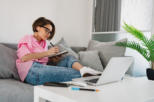 Attraktive, beschäftigte, ernste Frau in rosafarbenem Hemd, die konzentriert sitzt und Notizen macht, um Rechnungen auf dem Sofa zu Hause am Tisch zu bezahlen, der online am Laptop arbeitet