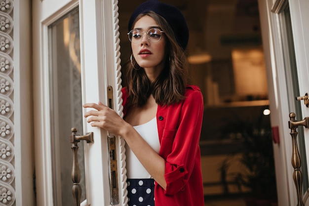 Attraktive blasse Frau mit klassischem Make-up-Tupfenrock, hellem, rotem Hemd und stilvollen Accessoires, die vor einem weißen und goldenen Vintage-Hintergrund posiert und wegschaut