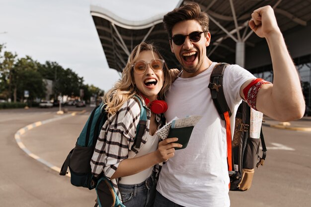 Attraktive blonde Frau mit Sonnenbrille und Mann in weißem T-Shirt lächelt und macht Selfie in der Nähe des Flughafens