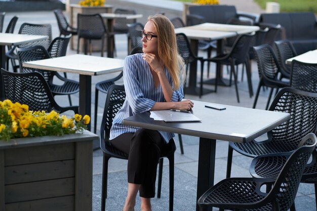 Attraktive blonde Geschäftsfrau denkt über ihren Job nach, während sie draußen in der Cafeteria mit Papierkram sitzt.