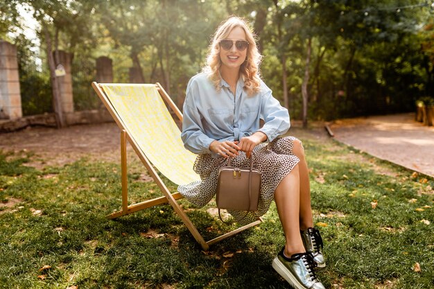 Attraktive blonde glückliche Frau, die entspannend im Liegestuhl im blauen Hemd des Sommeroutfits sitzt und silberne Turnschuhe, elegante Sonnenbrille und Geldbörse trägt