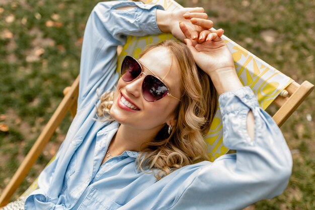 Attraktive blonde lächelnde Frau, die entspannt im Liegestuhl im stilvollen Outfit sitzt