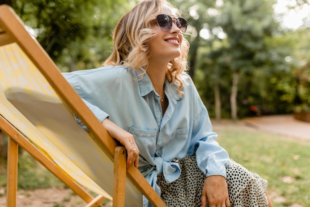 Attraktive blonde lächelnde Frau, die im Liegestuhl im Sommeroutfit sitzt