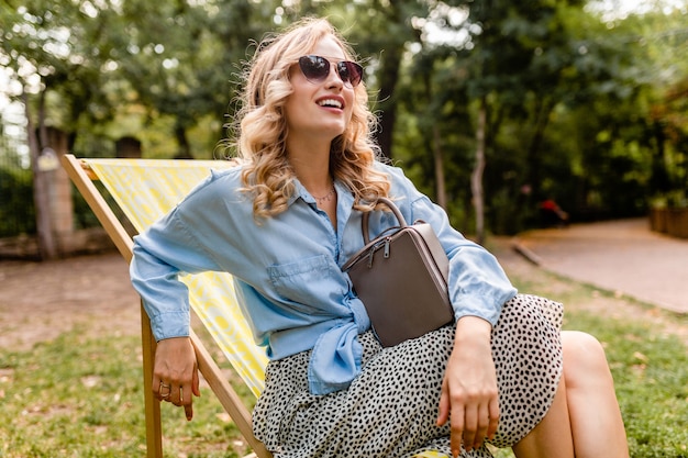 Attraktive blonde lächelnde Frau, die im Liegestuhl im Sommeroutfit sitzt