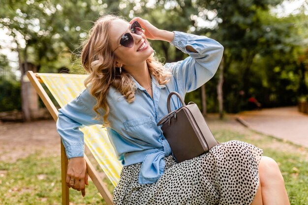 Attraktive blonde lächelnde Frau, die im Liegestuhl im Sommeroutfit sitzt