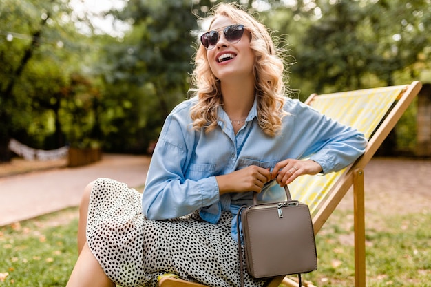 Attraktive blonde lächelnde Frau, die im Liegestuhl im Sommeroutfit sitzt
