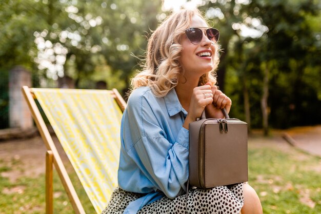 Attraktive blonde lächelnde Frau, die im Liegestuhl im Sommeroutfit sitzt