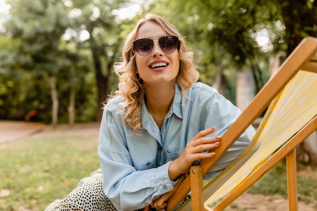 Attraktive blonde lächelnde Frau, die im Liegestuhl im stilvollen Outfit sitzt