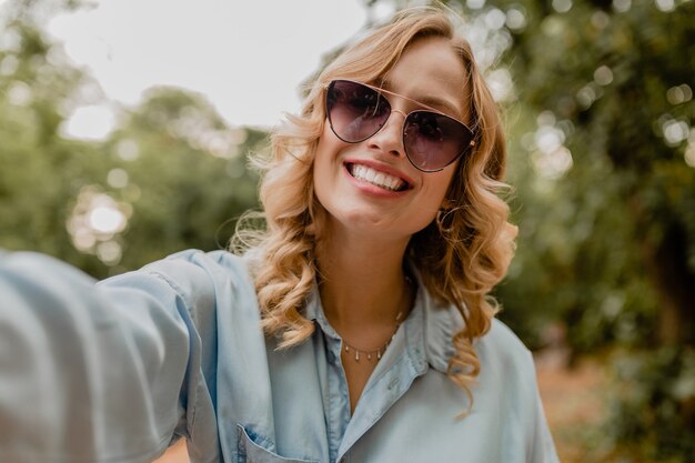 Attraktive blonde lächelnde Frau, die im Park im Sommeroutfit geht und Selfie-Foto am Telefon macht