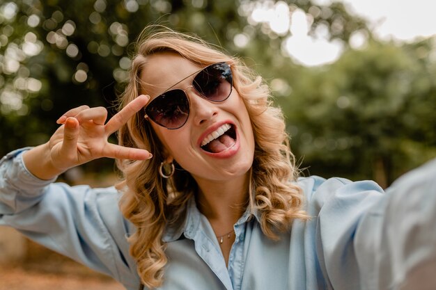 Attraktive blonde lächelnde Frau, die im Park im Sommeroutfit geht und Selfie-Foto am Telefon macht