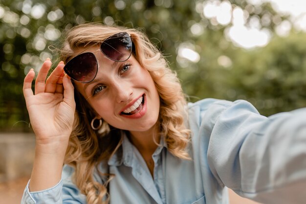 Attraktive blonde lächelnde Frau, die im Park im Sommeroutfit geht und Selfie-Foto am Telefon macht