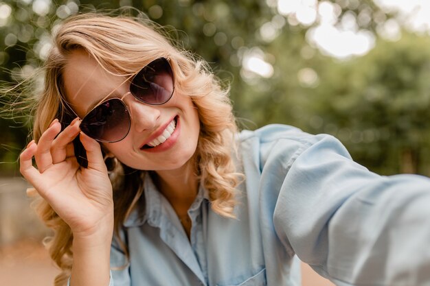 Attraktive blonde lächelnde Frau, die im Park im Sommeroutfit geht und Selfie-Foto am Telefon macht