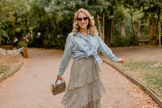 Attraktive blonde lächelnde Frau, die im Park im Sommeroutfit geht