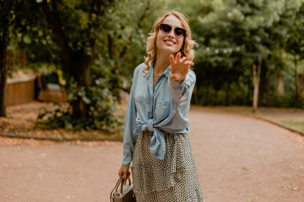 Attraktive blonde lächelnde Frau, die im Park im stilvollen Outfit geht