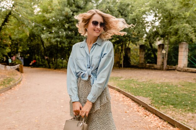 Attraktive blonde lächelnde offene Frau, die im Park im Sommeroutfit geht