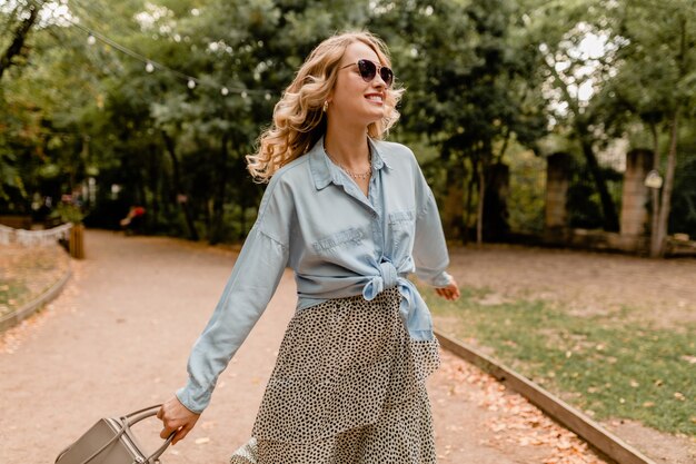 Attraktive blonde lächelnde offene Frau, die langes Haar winkt, das Spaß hat, im Park im Sommeroutfit zu gehen