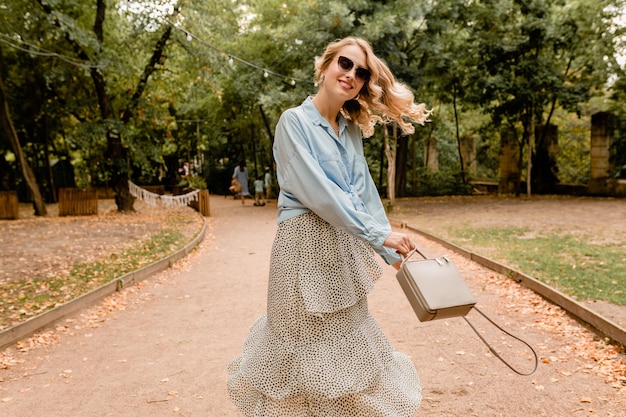 Attraktive blonde lächelnde offene Frau, die langes Haar winkt, das Spaß hat, im Park im Sommeroutfit zu gehen