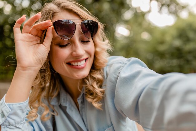 Attraktive blonde lächelnde weiße Zahnfrau, die im Park im Sommeroutfit geht, das selfie Foto am Telefon nimmt