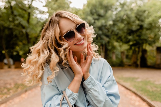 Attraktive blonde offene Frau, die im Park im stilvollen Outfit geht, das elegante Sonnenbrille und Geldbörse trägt