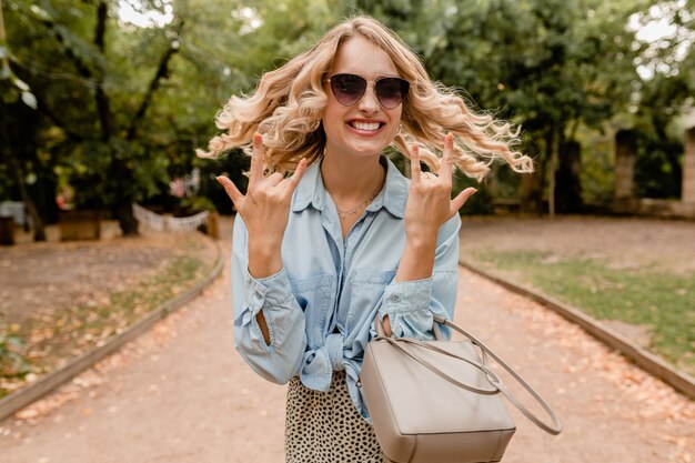 Attraktive blonde offene Frau, die im Park im stilvollen Outfit geht, das elegante Sonnenbrille und Geldbörse trägt