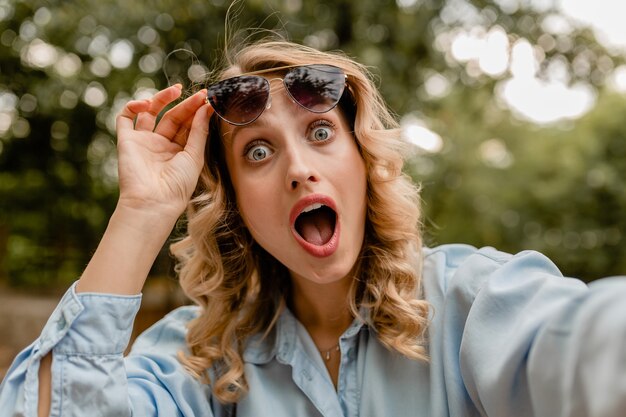 Attraktive blonde überraschte lustige Frau im Sommeroutfit, das Selfie-Foto am Telefon macht