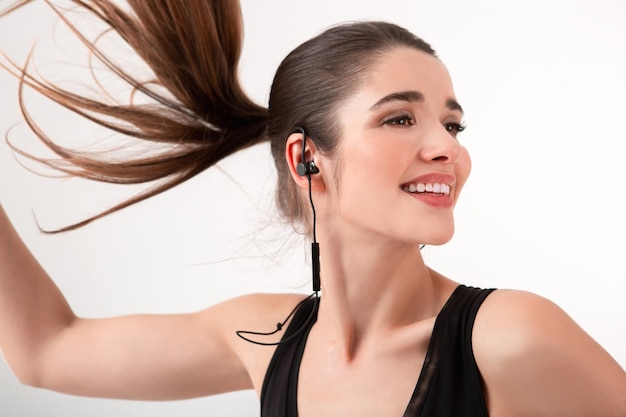 Attraktive brünette Frau in joggendem schwarzem Oberteil, die Musik über Kopfhörer hört und isoliert auf weißem Hintergrund posiert. Pferdeschwanzfrisur winkt mit langen Haaren