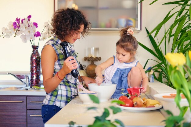 Attraktive brünette Frau mit lockigem Haar und ihrer süßen kleinen Tochter, die in einer Wohnküche Essen kocht.