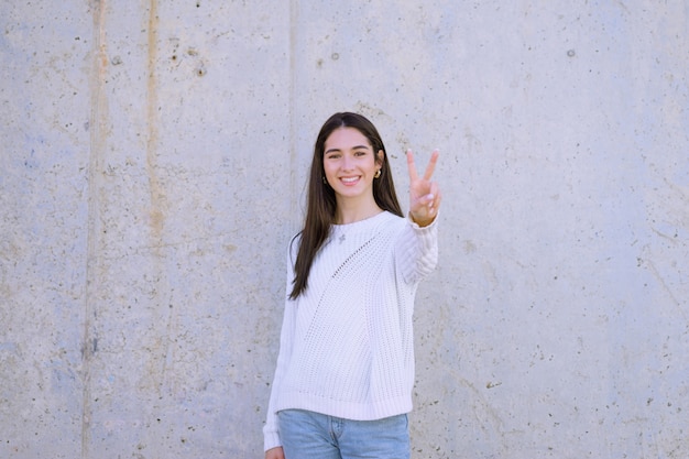 Attraktive Brünette mit einem glücklichen Lächeln, das einen weißen Pullover trägt, der das Friedenszeichen zeigt