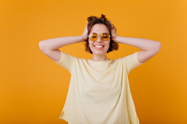 Attraktive Dame mit dunklem welligem Haar, das Glück ausdrückt. Innenporträt des glückseligen europäischen Mädchens in der gelben Kleidung lokalisiert auf orange Wand.