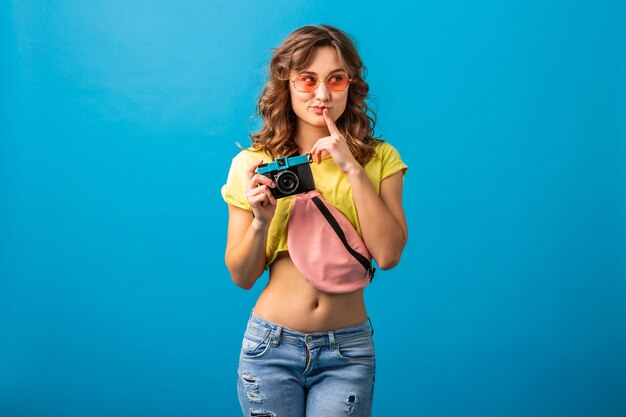 Attraktive denkende Frau, die mit Weinlesefoto-Kamera aufwirft, die Bilder macht, die im bunten Hipster-Sommeroutfit lokalisiert auf blauem Studiohintergrund gekleidet werden