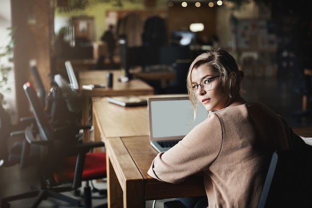 Attraktive entschlossene kaukasische blonde Frau in Brille, die sich umdreht, um in die Kamera zu schauen, von einem Kollegen angerufen wird, während er im Büro sitzt, über einen Laptop arbeitet und mit Kunden kommuniziert.
