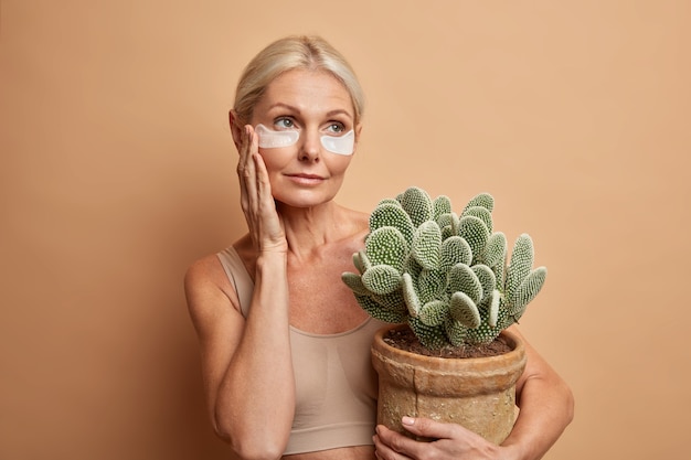 attraktive ernsthafte blonde alte Frau mit blonden Haaren berührt Gesicht trägt Schönheitsflecken unter den Augen umarmt Topf Kaktus