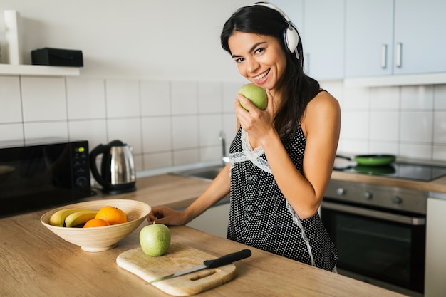 Attraktive Frau der jungen Brünette, die in der Küche am Morgen kocht, grünen Apfel isst, lächelt, glückliche Stimmung, positive Hausfrau, gesunden Lebensstil, Musik auf Kopfhörern hört