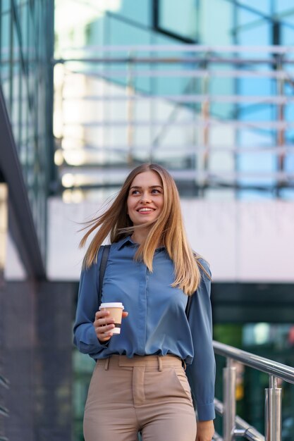 Attraktive Frau der Nahaufnahme in Bewegung mit Kaffee zum Mitnehmen am Geschäftsgebäude. Porträt blondes Mädchen, das Pappbecher mit heißem Getränk im Freien hält.