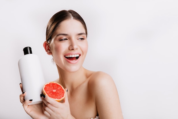 Attraktive Frau, die freundlich auf weißer Wand lächelt. Mädchen ohne Make-up zeigt Trauben- und Haarshampoo.