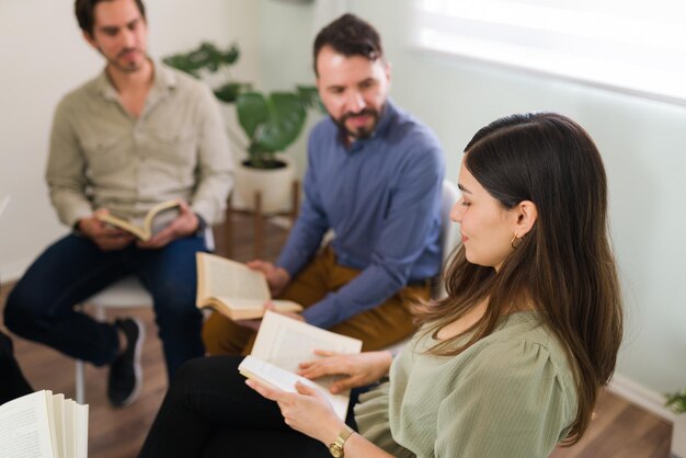 Attraktive Frau, die lächelt, während sie während eines Treffens ihres Buchclubs mit jungen Leuten ein Zitat aus einem Buch laut vorliest