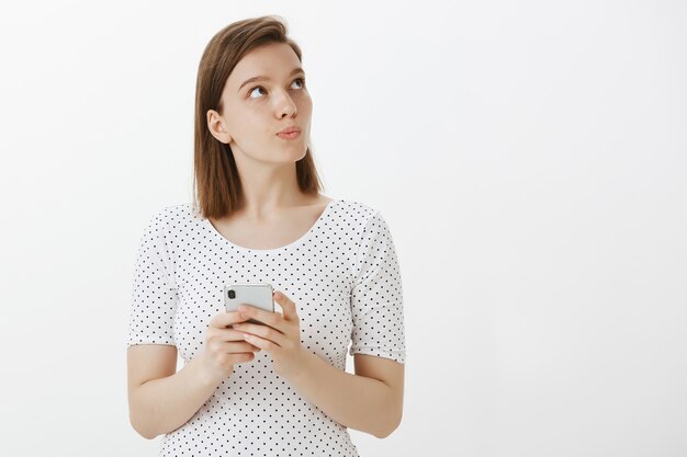 Attraktive Frau, die nachdenkliche obere rechte Ecke schaut, während sie denkt, wie auf Nachricht zu antworten, Handy hält