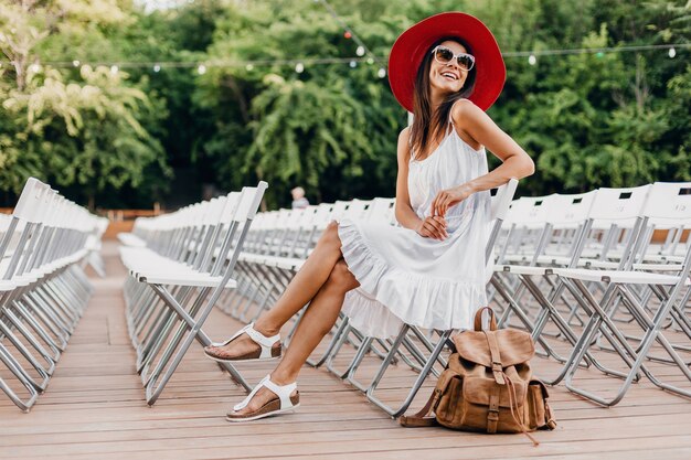 Attraktive Frau gekleidet in weißem Kleid, rotem Hut, Sonnenbrille, die im Sommer Freilufttheater allein auf Stuhl sitzt, Modetrend im Frühlingsstraßenstil, Accessoires, Rucksack, soziale Distanzierung