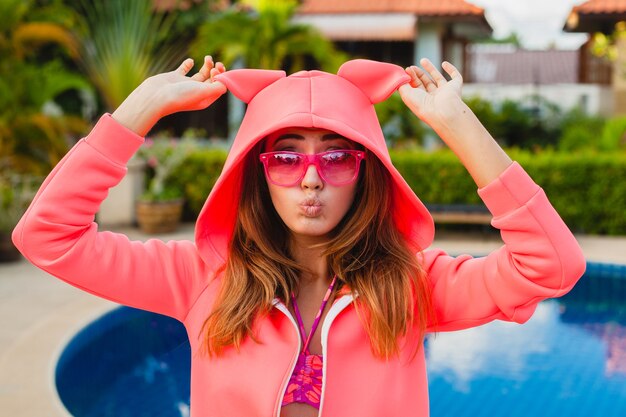 Attraktive Frau im bunten rosa Kapuzenpulli, der Sonnenbrillen auf Sommerferien lächelndem emotionalen Gesichtsausdruck trägt, der Spaß, Sportmode-Stil hat