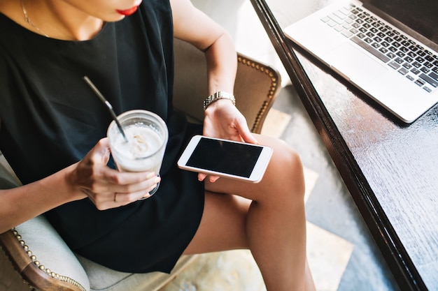 Attraktive Frau im kurzen schwarzen Kleid im Stuhl, Telefon und Glas Cappuccino haltend.